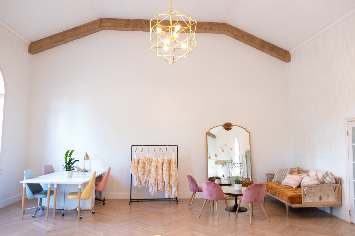 inside the pleiades studio. There are big wooden beams, dresses on racks, a big antique mirror, and pastel colored chairs.