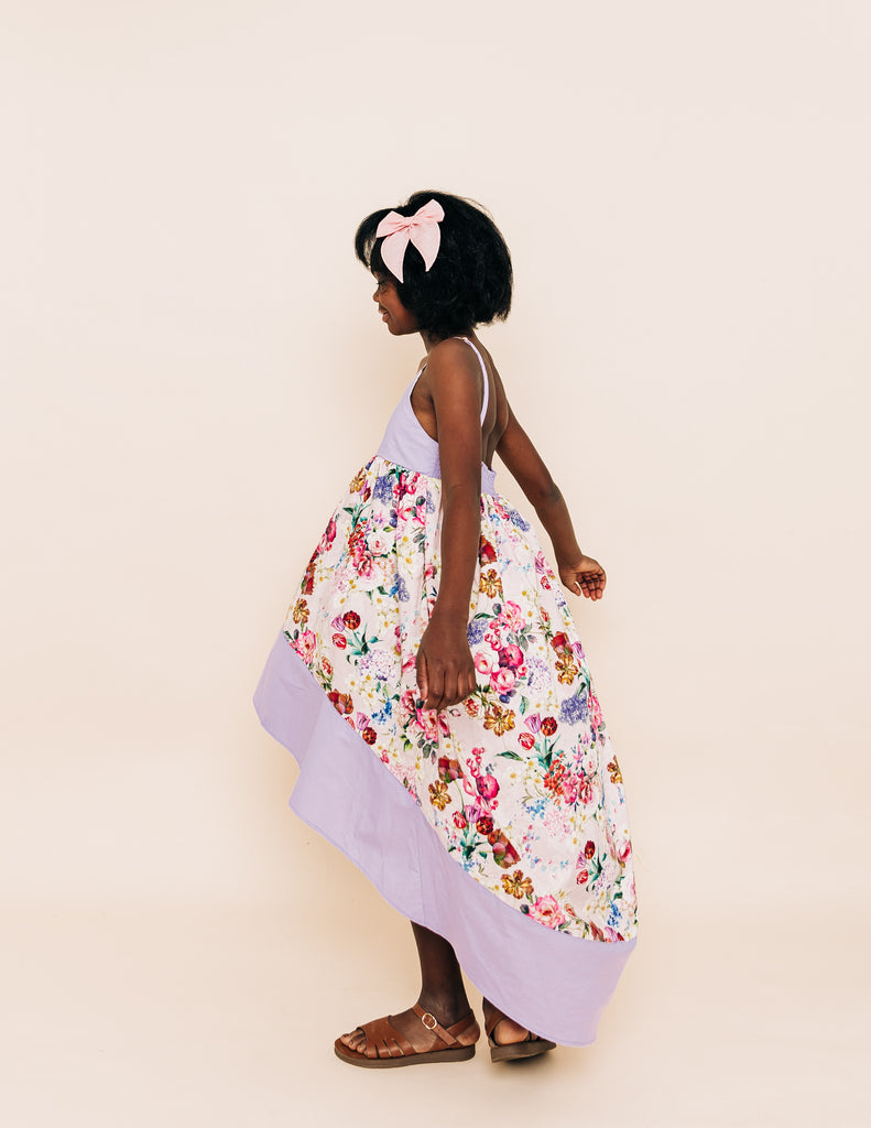 Girl wearing a floral high low dress that has a purple hem and bodice. Accessorized with a pink bow in her hair