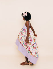 Girl wearing a floral high low dress that has a purple hem and bodice. Accessorized with a pink bow in her hair