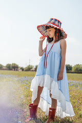 Girl in a blue flower field wearing a blue clip dot high low dress with a white hem. Accessorized with red boots and a red and blue hat