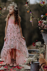 Girl surrounded by flowers wearing a flower crown and a pink Pleiades Designs high low dress with embroidered rosebuds and a ruffle hem